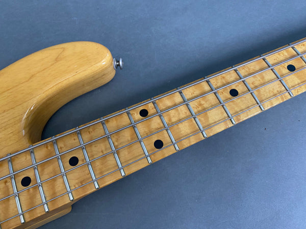 Close-up of maple fingerboard and neck on natural wood electric bass guitar with black dot inlays and metal frets against gray background