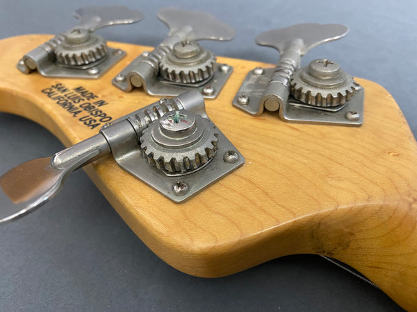 Close-up view of vintage bass guitar tuning pegs on natural wood headstock with "Made in San Luis Obispo California USA" stamp, silver tuning machines, grey background