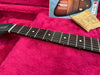 Close-up of guitar neck and fretboard with dot inlays resting on a pink plush guitar case lining with vintage guitar magazine and cleaning cloth in the background.