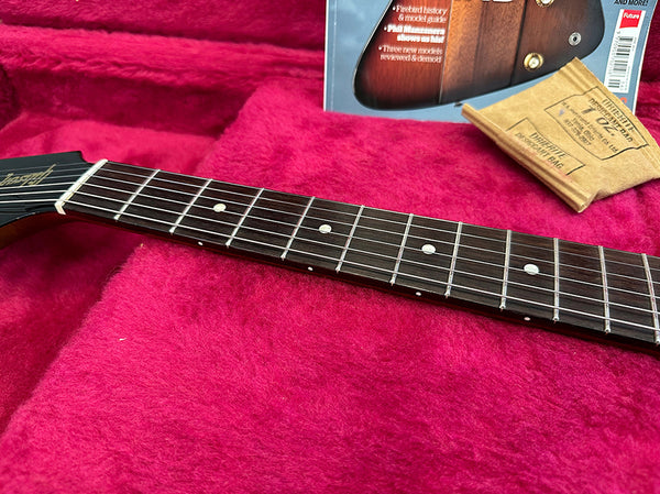 Close-up of guitar neck and fretboard with dot inlays resting on a pink plush guitar case lining with vintage guitar magazine and cleaning cloth in the background.