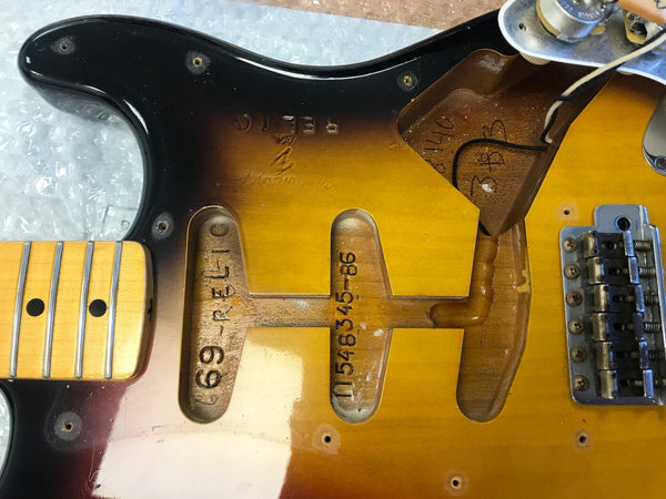 Close-up of disassembled vintage electric guitar body showing pickup cavities with handwritten serial numbers and "69-RELIC" marking, maple neck with black dot inlays, and metal bridge hardware on sunburst finish guitar body.