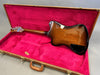Electric guitar with sunburst finish inside open vintage brown hard case lined with red plush interior.