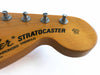 Close-up of Fender Stratocaster guitar headstock with tuning pegs and black lettering on natural wood finish, featuring original contour body text