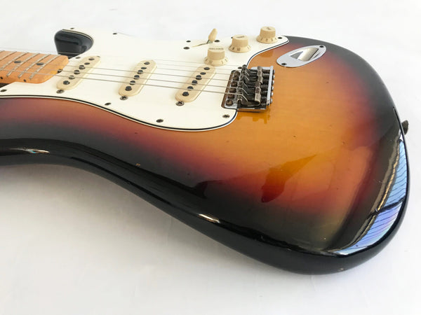 Close-up of sunburst electric guitar body with white pickguard, three single-coil pickups, and volume and tone control knobs on white background