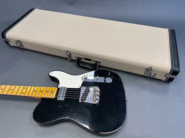 Black electric guitar with maple neck and white pickguard next to beige hard guitar case on gray background