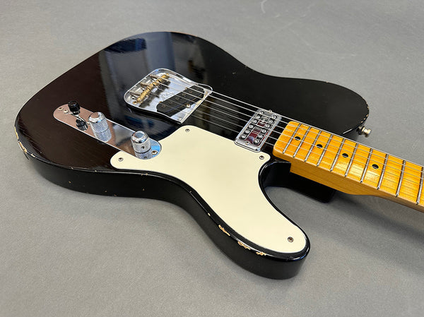 Black electric guitar with maple neck and white pickguard, showing worn edges and chrome hardware, laid on gray surface