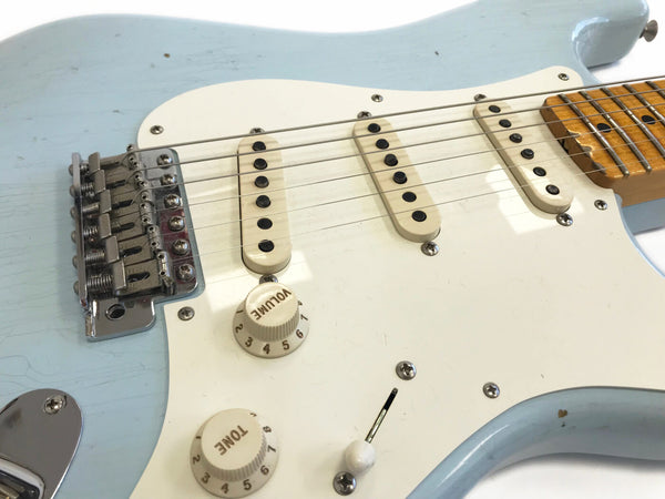 Close-up of light blue electric guitar body showing white pickguard, three single-coil pickups, volume and tone control knobs, and chrome bridge with six saddles.