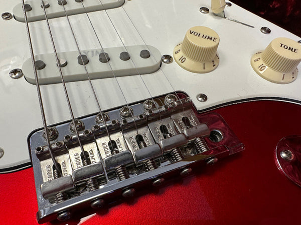 Close-up of red electric guitar bridge, white pickguard, volume and tone knobs, and single coil pickups with metal saddles and strings at Coffee House Guitars
