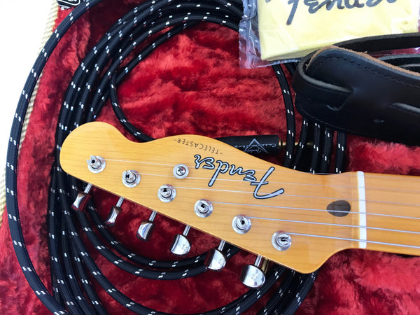Close-up of Fender Telecaster guitar headstock with chrome tuning pegs, black braided Fender guitar cable, black leather strap, and yellow Fender polishing cloth on red plush surface
