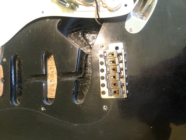 Close-up of black Fender Stratocaster electric guitar body showing empty pickup cavities, serial number stamp inside cavity, and vintage-style bridge with six adjustable saddles and "Fender" engravings on saddles