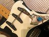 Close-up of a black electric guitar with the pickguard removed, showing three single-coil pickups and wiring on a white pickguard, with exposed potentiometers and capacitor components.