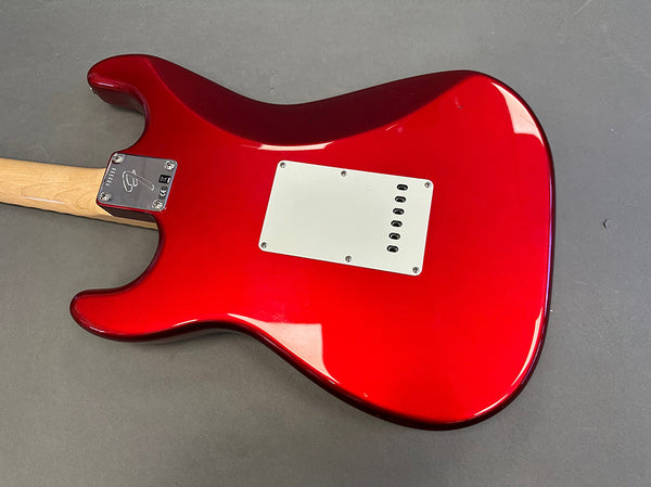 Close-up of the back of a red electric guitar body with a white backplate and maple neck on a gray surface, showcasing the guitar's sleek finish and chrome neck plate with guitar logo.