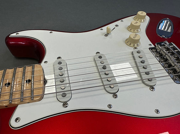 Close-up of red electric guitar body with white pickguard, three single-coil pickups, maple fretboard, and cream tone and volume knobs.
