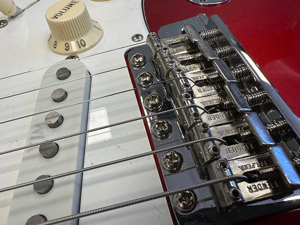 Close-up of red electric guitar bridge with Fender saddles and white volume knob on white pickguard with single-coil pickups and steel guitar strings