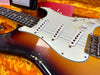 Close-up of vintage sunburst electric guitar body with white pickguard, three single-coil pickups, volume and tone knobs, and worn finish on orange plush case lining