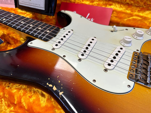 Close-up of vintage sunburst electric guitar body with white pickguard, three single-coil pickups, volume and tone knobs, and worn finish on orange plush case lining