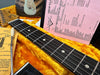 Close-up of Fender Stratocaster guitar neck and fretboard with dot inlays, tremolo arm, vintage-style tuning instructions, and pink Fender shop setup sheet inside a yellow plush guitar case.