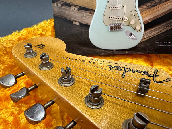 Close-up of vintage Fender Stratocaster guitar headstock with tuning pegs on orange plush case interior and faded surf green guitar in background