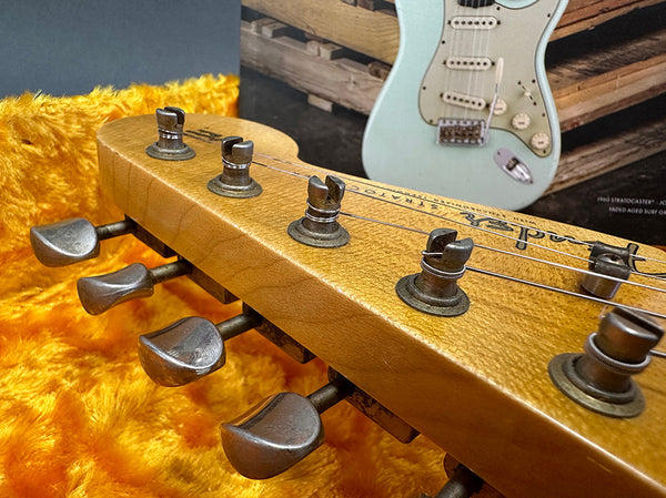 Close-up of vintage Fender Stratocaster guitar headstock with worn tuning pegs and strings, resting on orange plush fabric with a Stratocaster guitar poster in background.