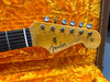 Close-up of Fender Stratocaster headstock with six tuning pegs, natural wood finish, and original contour body logo, displayed on bright orange plush guitar case interior.