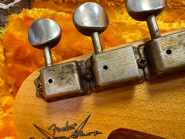 Close-up of vintage Fender Custom Shop guitar tuning pegs with aged metal finish on light wood headstock against orange plush background