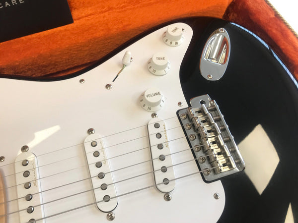 Close-up of black electric guitar with white pickguard showing tone and volume control knobs, three single-coil pickups, and chrome bridge in orange-lined case