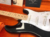 Close-up of black Fender electric guitar with white pickguard and maple neck resting in orange plush-lined case with Fender Custom Shop documentation visible in the background.