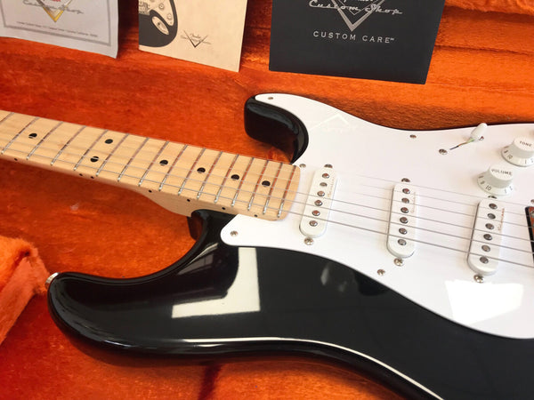 Close-up of black Fender electric guitar with white pickguard and maple neck resting in orange plush-lined case with Fender Custom Shop documentation visible in the background.