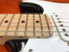 Close-up of maple guitar neck with metal frets, black dot fret markers, and steel strings on a white electric guitar body against an orange background.