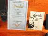 Fender Certificate of Authenticity for Clapton Stratocaster with serial number CZ524639 and Fender Lacquer Finishes Precautions and Care booklet on orange guitar case interior next to black Fender Custom Shop leather strap.