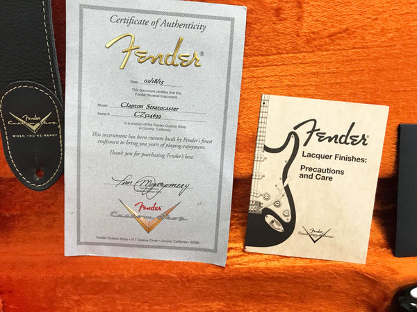 Fender Certificate of Authenticity for Clapton Stratocaster with serial number CZ524639 and Fender Lacquer Finishes Precautions and Care booklet on orange guitar case interior next to black Fender Custom Shop leather strap.