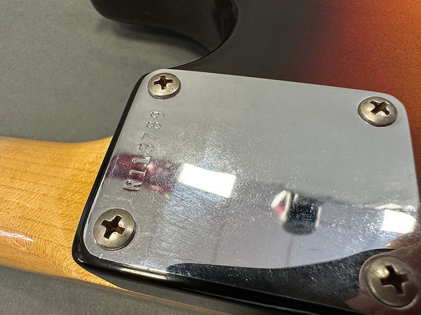 Close-up of metal neck plate on electric guitar with serial number R113789 and four screws, showing wood grain of guitar neck and sunburst finish on body.