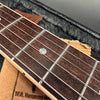 Close-up of guitar fretboard with metal frets and pearl dot inlay, steel strings visible, dark wood neck, displayed in black padded guitar case with cardboard underneath.