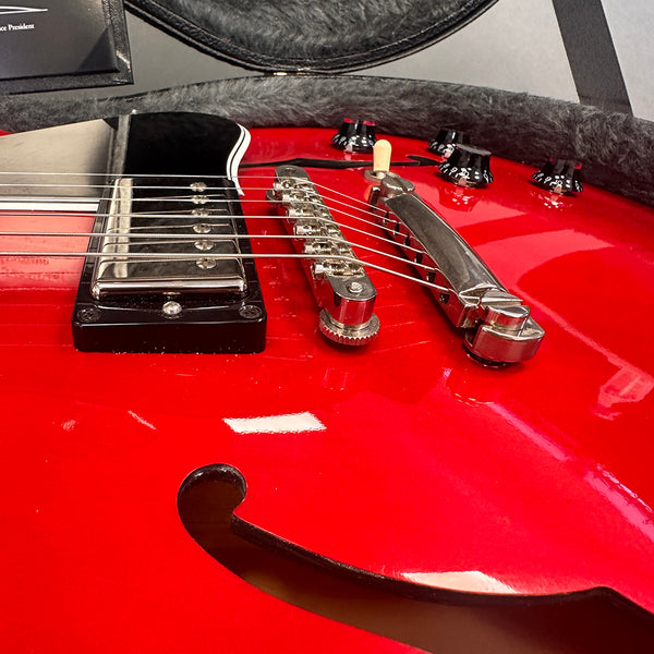 Close-up of red semi-hollow electric guitar body showing chrome bridge, tailpiece, black pickup, control knobs, and f-hole design in guitar case lining