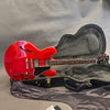 Red semi-hollow electric guitar with dual humbucker pickups, black pickguard, and chrome hardware resting in an open black guitar case with plush grey interior lining on a grey background.