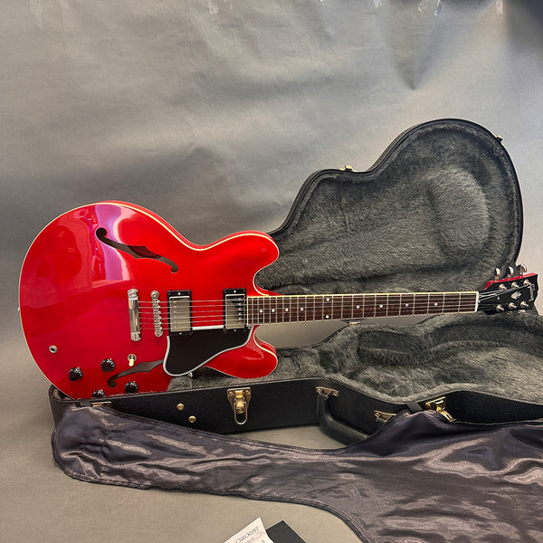 Red semi-hollow electric guitar with dual humbucker pickups, black pickguard, and chrome hardware resting in an open black guitar case with plush grey interior lining on a grey background.