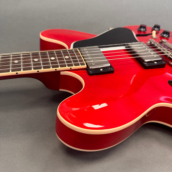 Close-up of red semi-hollow electric guitar body with cream binding and black pickguard on gray background, showing pickups and fretboard details