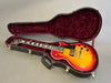 Electric guitar with sunburst finish in black hard case with red velvet interior lining, angled view showing body, neck, and headstock
