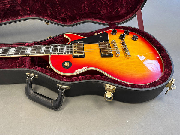 Sunburst electric guitar with black pickguard and gold hardware in black hardshell case with red velvet interior on gray surface