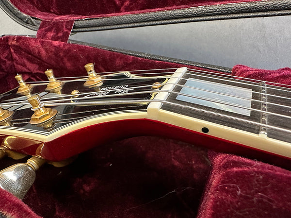 Close-up of black and red Les Paul Custom electric guitar headstock with gold tuning pegs and strings, resting in red velvet guitar case interior