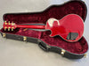 Red electric guitar with gold tuning pegs inside a black hard case with plush red interior, showing the back of the guitar and neck.