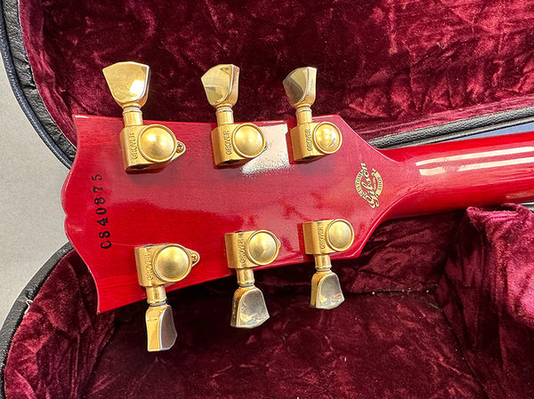 Close-up of red guitar headstock with gold Grover tuning pegs and Gibson Custom Shop insignia, inside a plush red velvet guitar case.