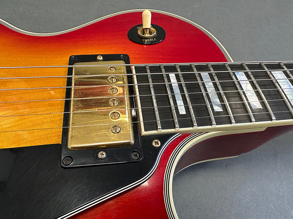 Close-up of electric guitar body showing sunburst finish, gold humbucker pickup, black pickguard, and white toggle switch on grey background