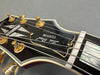 Close-up of black Les Paul Custom guitar headstock with gold tuning pegs and white binding on fretboard from Coffee House Guitars product listing.