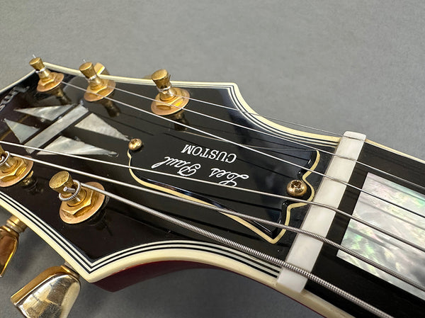 Close-up of black Les Paul Custom guitar headstock with gold tuning pegs and white binding on fretboard from Coffee House Guitars product listing.