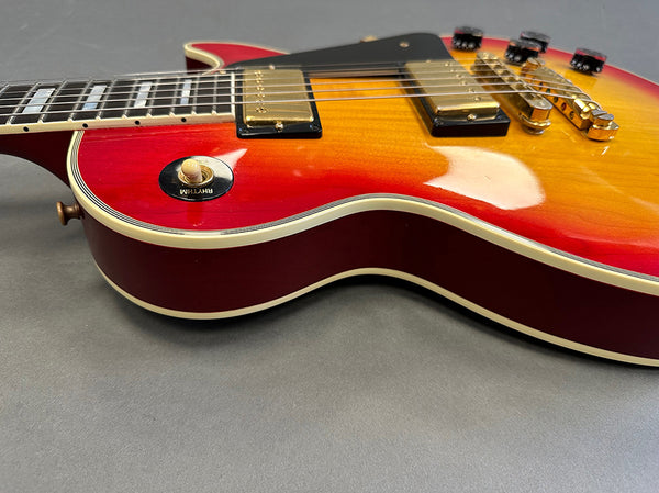 Close-up of sunburst electric guitar body showing rhythm switch, gold pickups, bridge, and strings on gray background