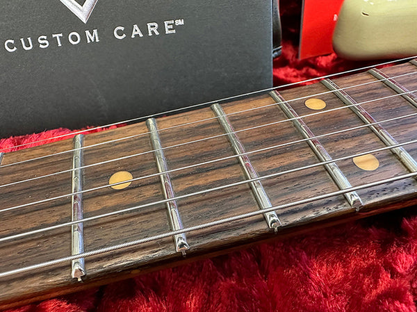 Close-up of guitar fretboard with nickel strings and dot inlays on a rosewood fingerboard inside a red plush guitar case with Custom Care logo visible