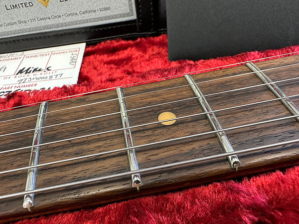 Close-up of guitar fretboard with metal frets and dot inlay on rosewood, displayed inside red velvet-lined guitar case with inspection and authenticity certificates visible in background.