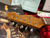 Close-up of vintage Fender Stratocaster guitar headstock with tuning pegs, Fender Custom Shop paperwork, and certification tags on a red velvet background, highlighting authenticity and craftsmanship.
