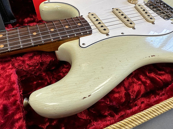 Close-up of vintage white electric guitar body with visible wear and cracks, rosewood fingerboard with dot inlays, and three single-coil pickups resting on red plush guitar case lining